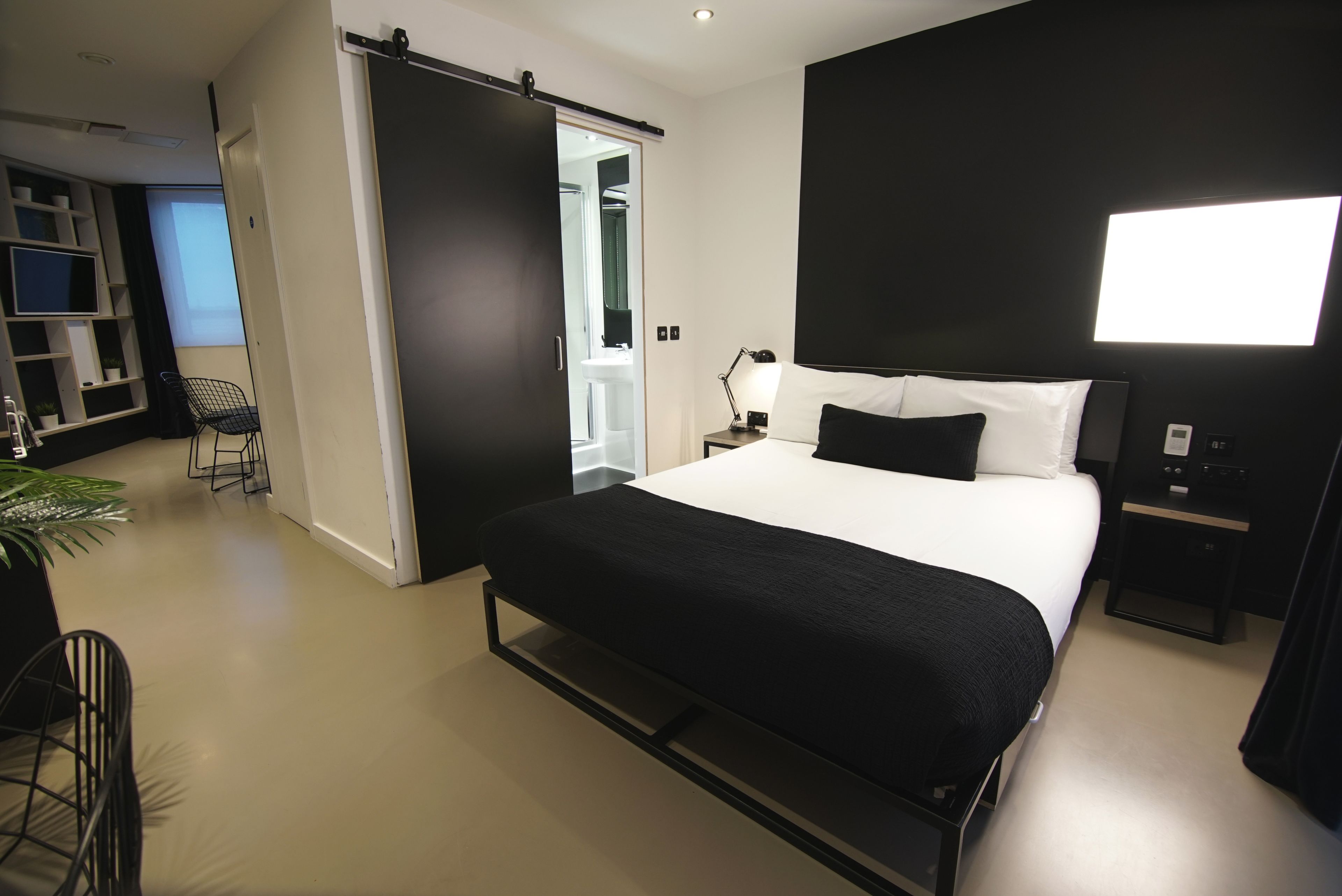 Moderni hotellihuone, jossa on suuri sänky, yöpöytä lampun kanssa, musta tuoli ja liukuovi, joka johtaa kylpyhuoneeseen.