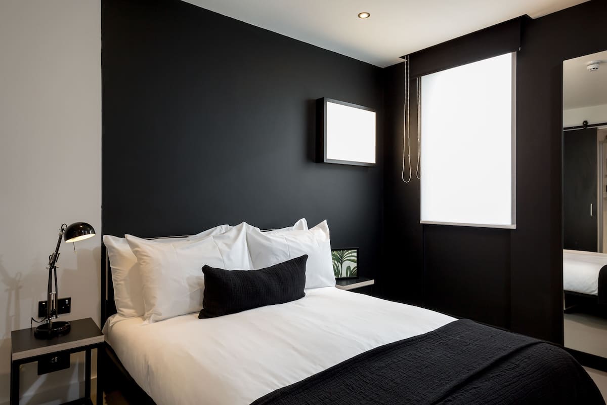 A modern bedroom with a large bed, a bedside table with a lamp, a framed picture on the wall, and a window with a view.