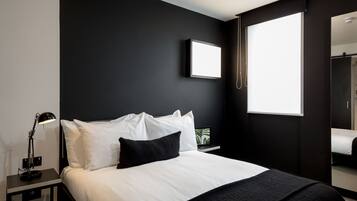 A modern bedroom with a large bed, a bedside table with a lamp, a framed picture on the wall, and a window with a view.