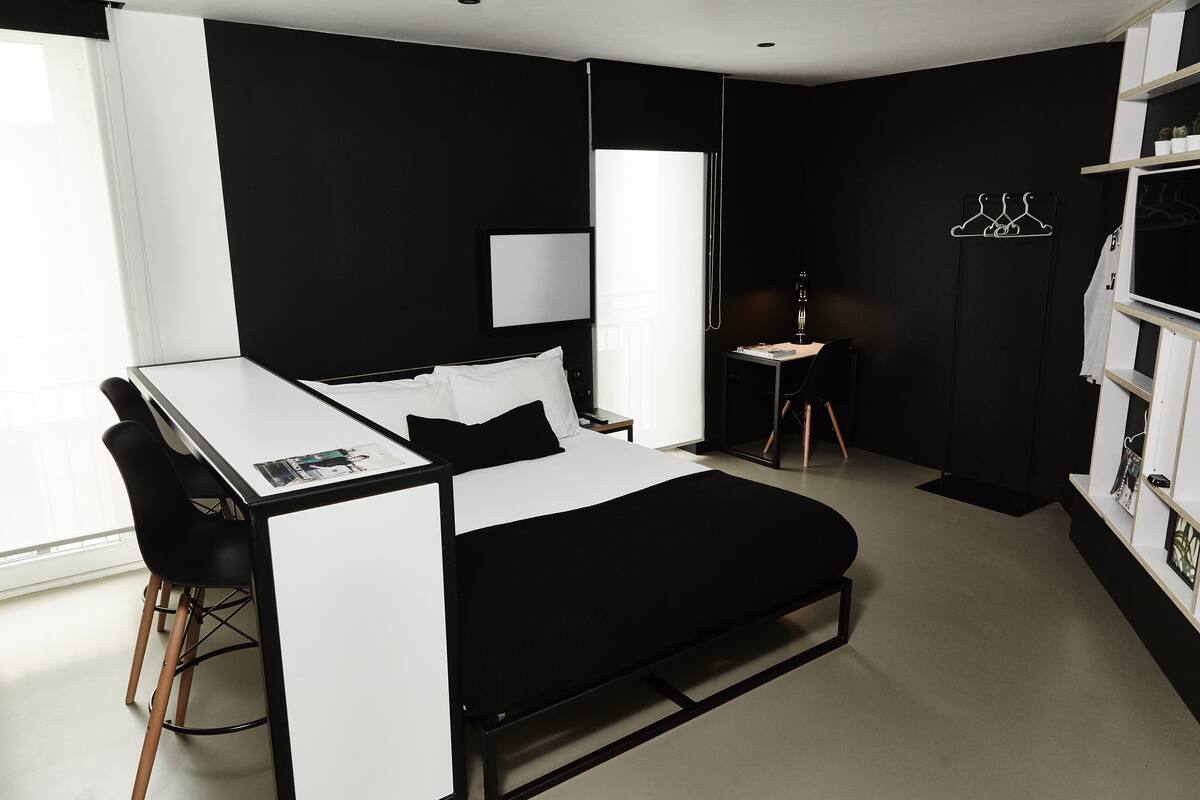 A modern hotel room with a bed, a desk, and a chair. There is a wardrobe with a mirror and a coat rack.