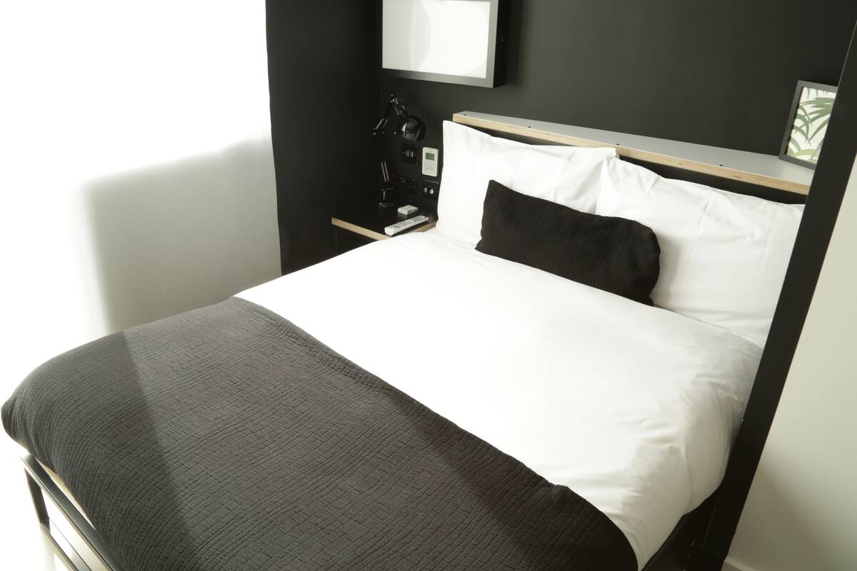 A neatly made bed with a white and gray duvet, a black pillow, and a bedside table with a lamp and a digital clock.