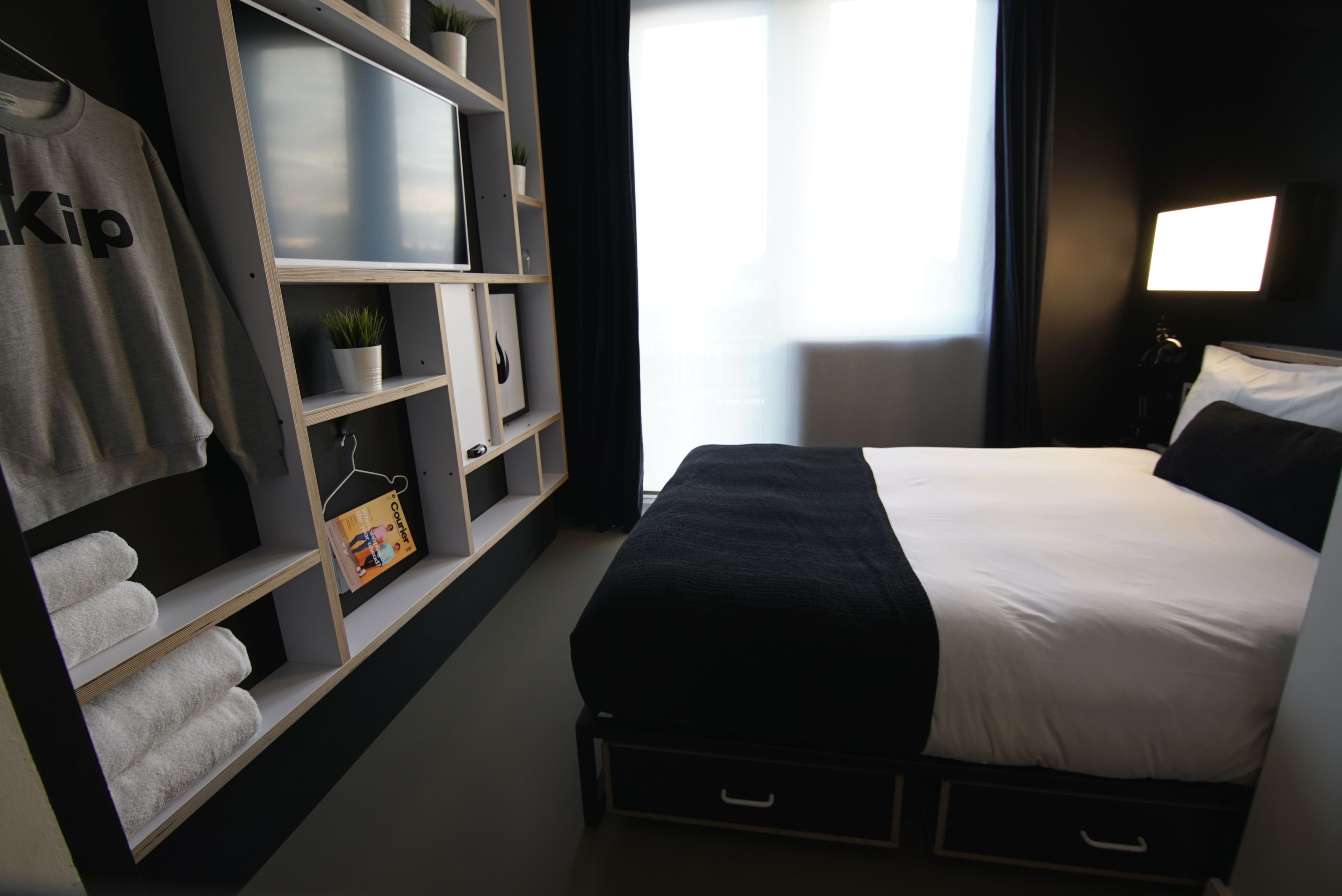 Une chambre d’hôtel bien rangée, avec un lit, une armoire dotée d’étagères, un porte-serviettes et une table de chevet avec une lampe.