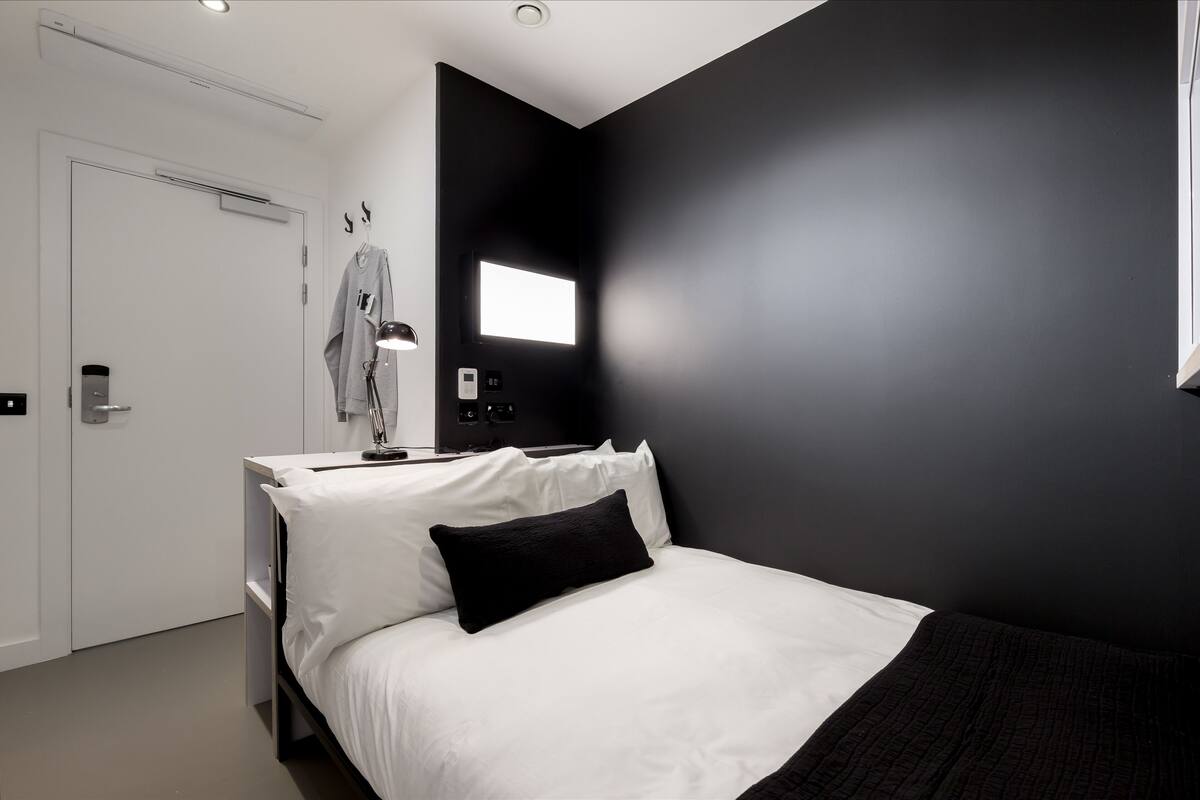 A modern bedroom with a bed, a desk, and a chair. There is a door and a window.