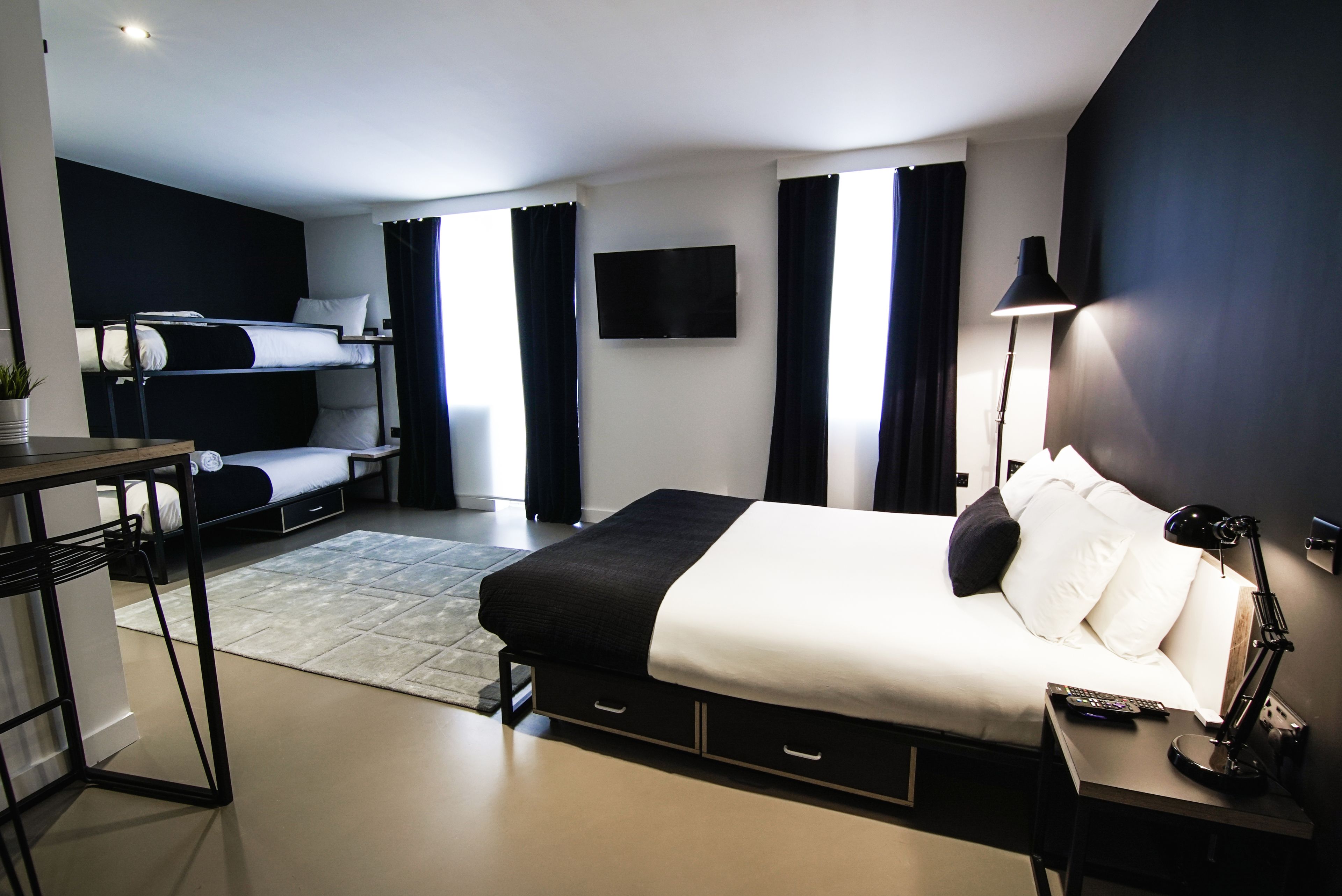 A modern hotel room with a bunk bed, a desk with a chair, a flat-screen TV, a desk lamp, and a large window with curtains.