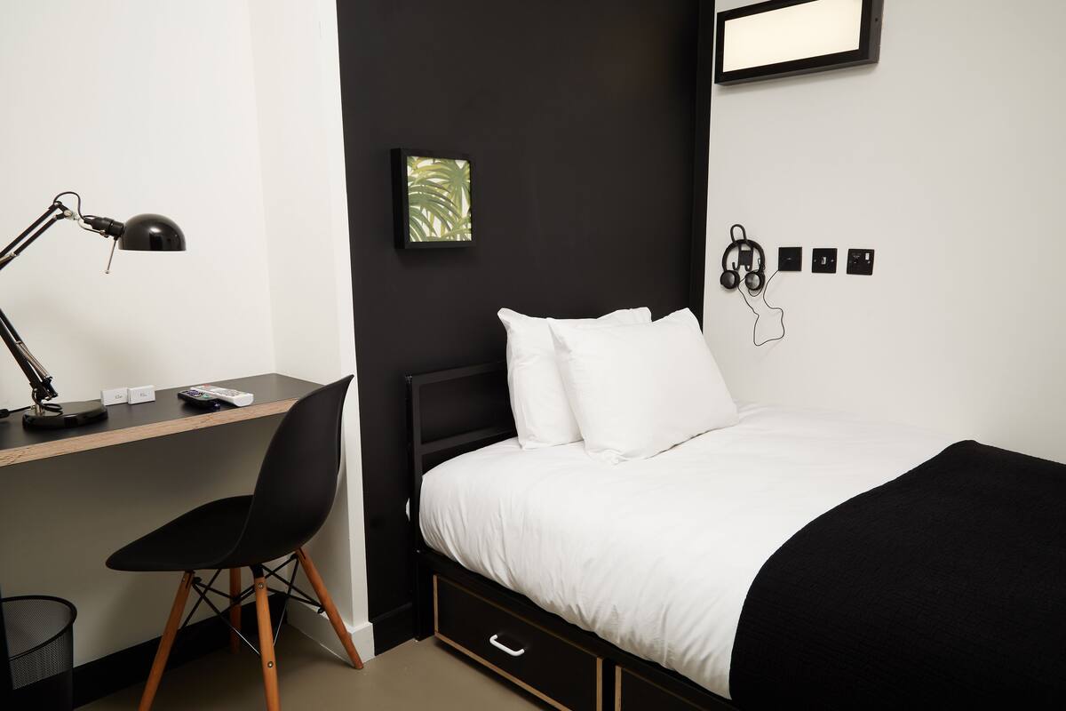 A modern bedroom with a bed, desk, chair, and a wall-mounted power outlet.