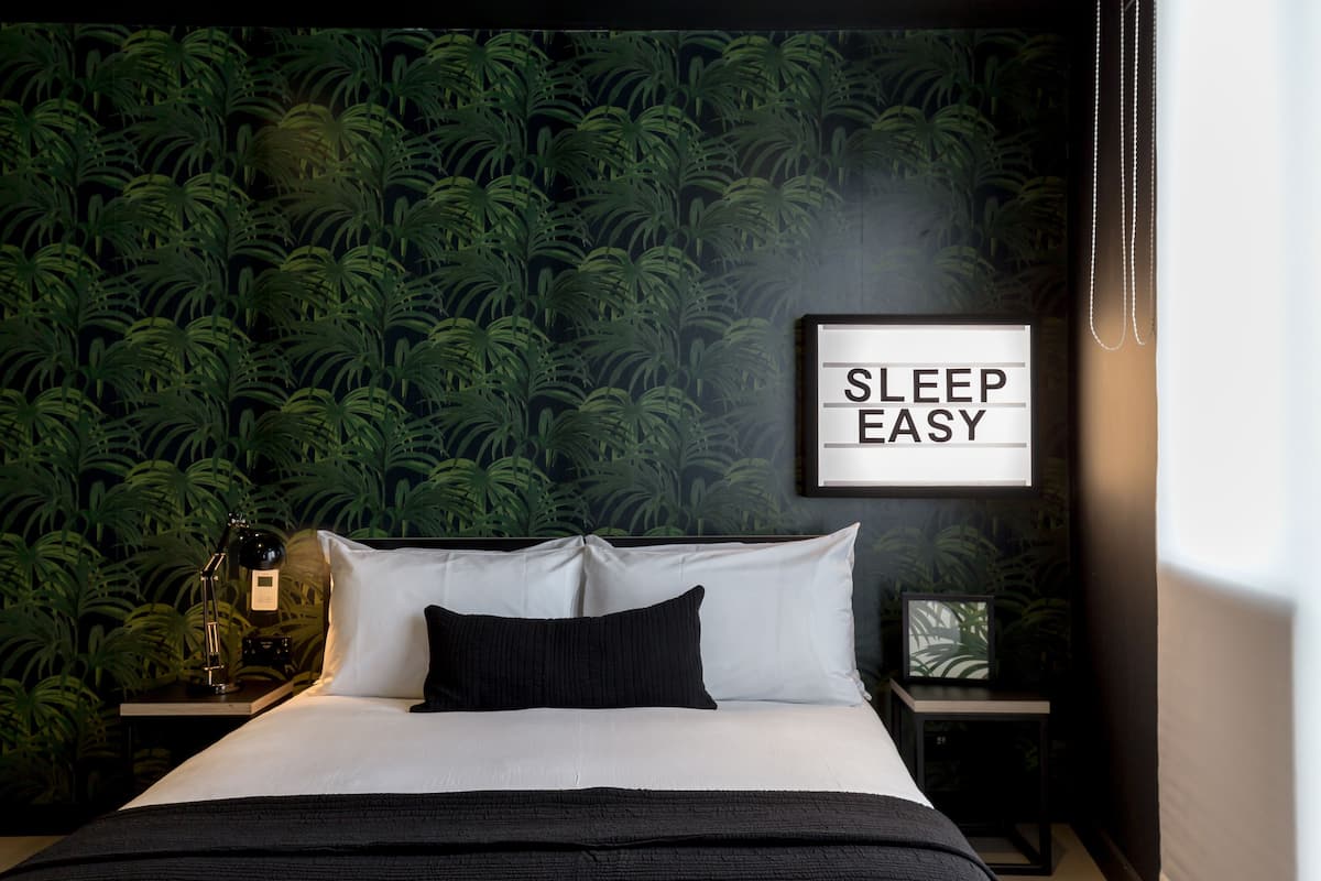 A neatly made bed with white and black bedding, a framed sign saying "SLEEP EASY" on the wall, and a nightstand with a lamp.
