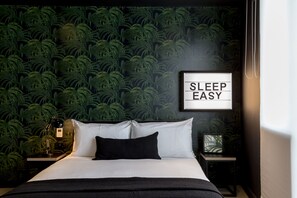 A neatly made bed with white and black bedding, a framed sign saying "SLEEP EASY" on the wall, and a nightstand with a lamp. - Kip Hotel (London)