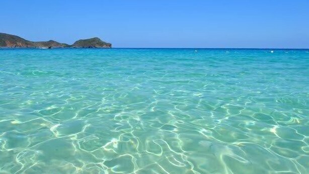 Helder turquoise water met zichtbare zandpatronen en verafgelegen rotsformaties.