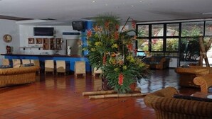 Een ruime lobby met een receptiebalie, rieten meubels en een grote kamerplant.