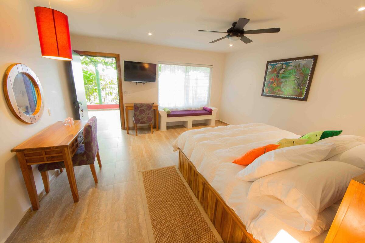 A bedroom with a bed, a desk, a chair, a television, and a window with a view of greenery.