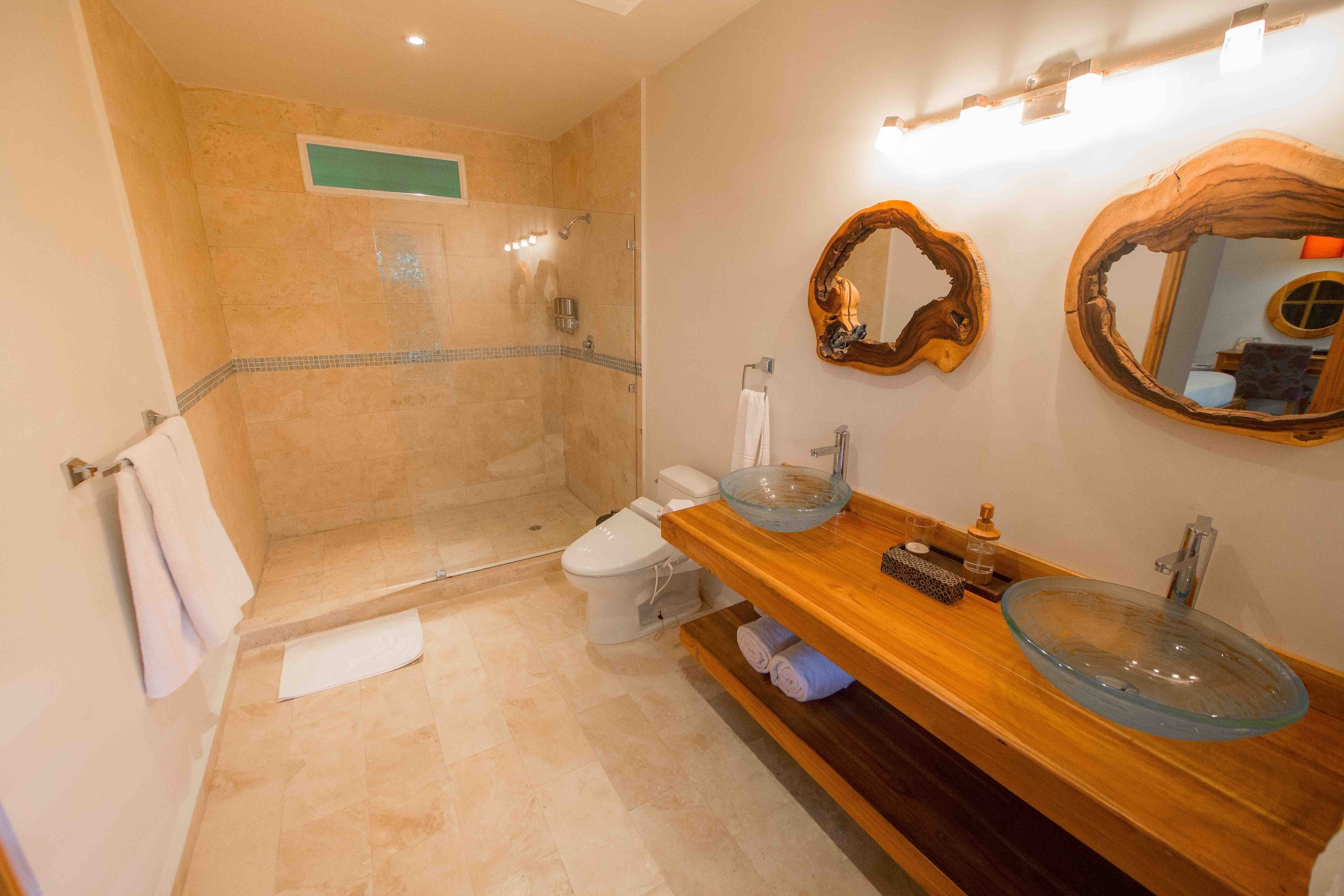 Un baño con lavabo de vidrio, tocador de madera, inodoro y ducha a pie.