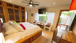 Un dormitorio con cama, escritorio, silla, televisor y balcón con vistas a la vegetación.