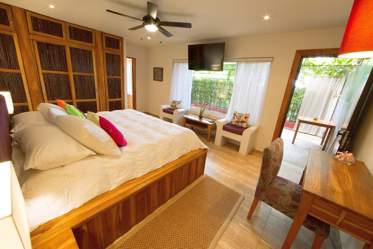 A bedroom with a bed, a desk, a chair, a TV, and a balcony with a view of greenery.