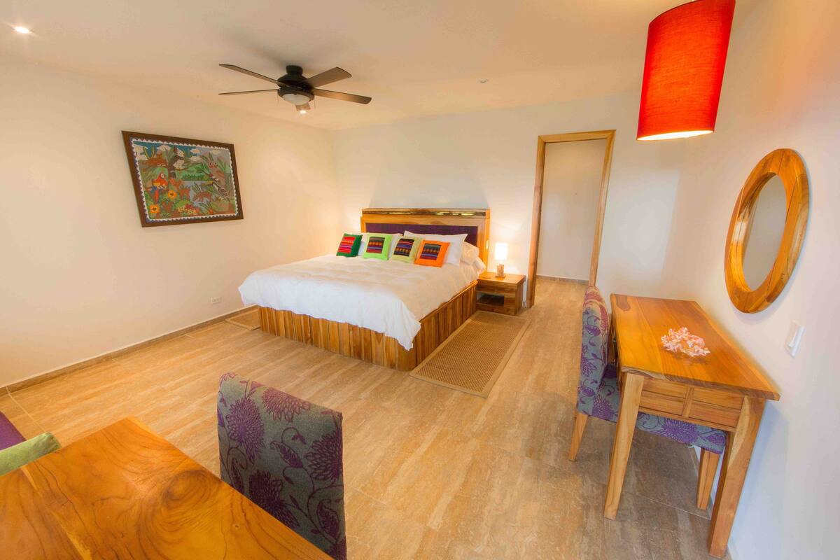 A hotel room with a bed, a desk, a chair, a ceiling fan, a wall-mounted mirror, and a floor lamp.