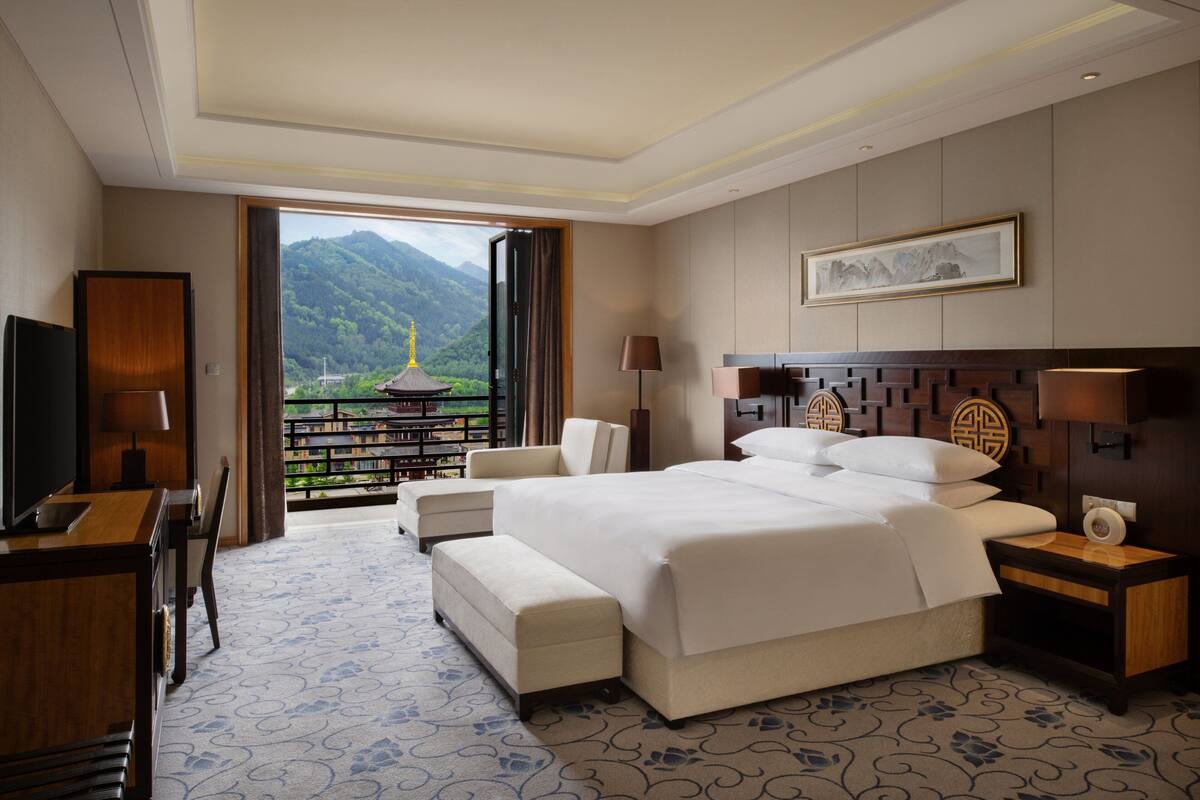 A hotel room with a large bed, a desk, a chair, and a view of mountains through an open door.