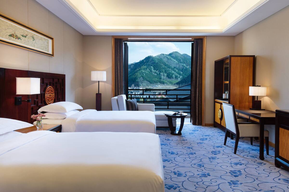 A hotel room with two beds, a desk, a chair, and a view of mountains.