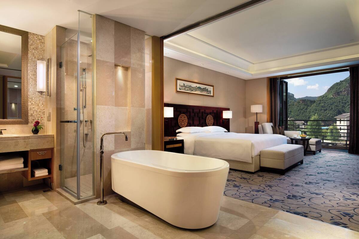 A modern hotel room with a large bed, a separate bathroom with a bathtub, a glass-enclosed shower, and a view of a mountainous landscape.