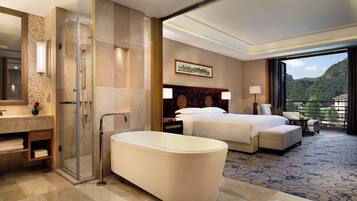 A modern hotel room with a large bed, a separate bathroom with a bathtub, a glass-enclosed shower, and a view of a mountainous landscape.
