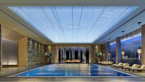 Un área de piscina interior moderna con techo de vidrio, paredes de piedra y una zona de estar con tumbonas.