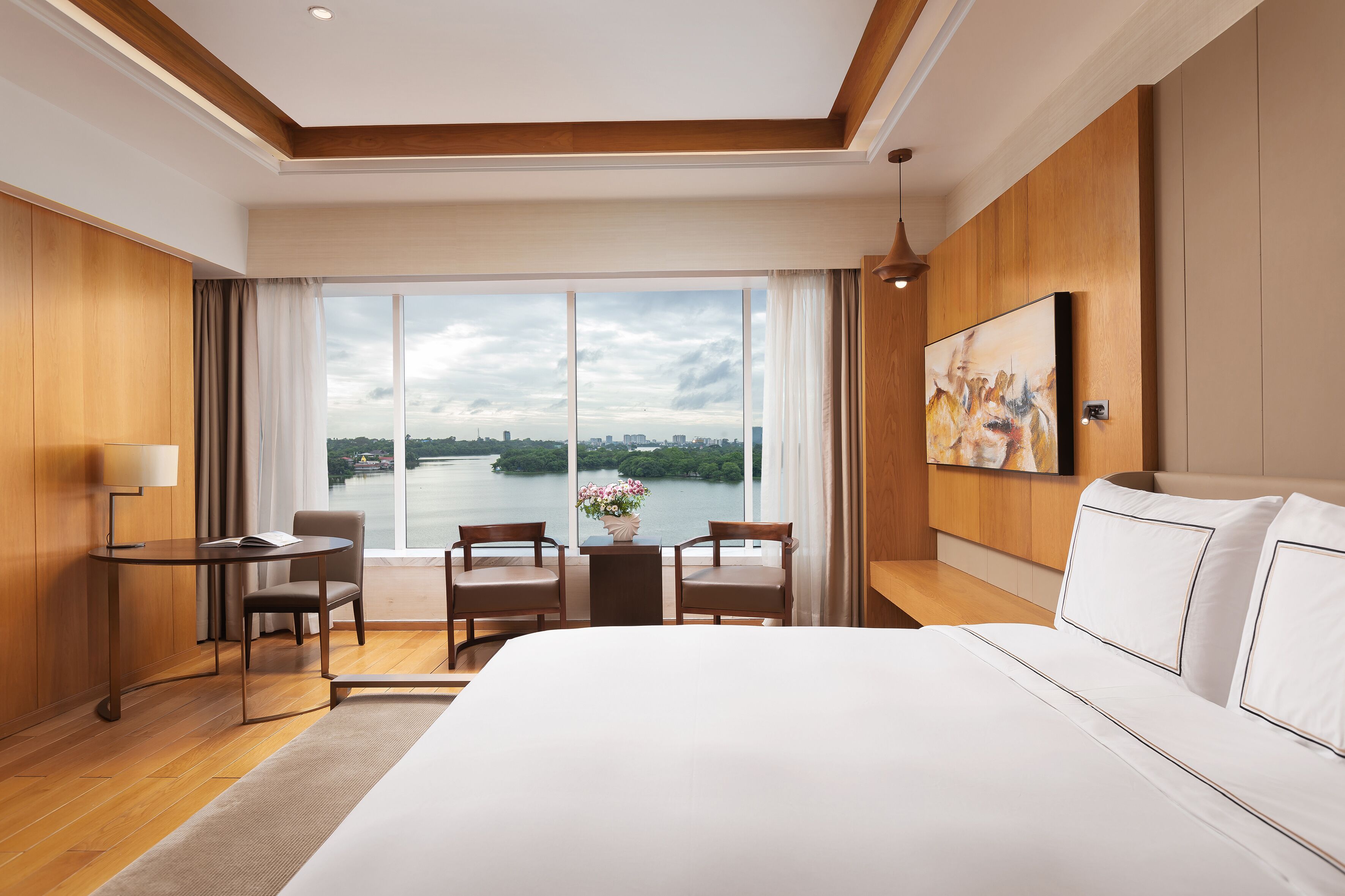 A hotel room with a large bed, a desk with a chair, a small table with a chair, and a large window offering a view of a body of water and cityscape.