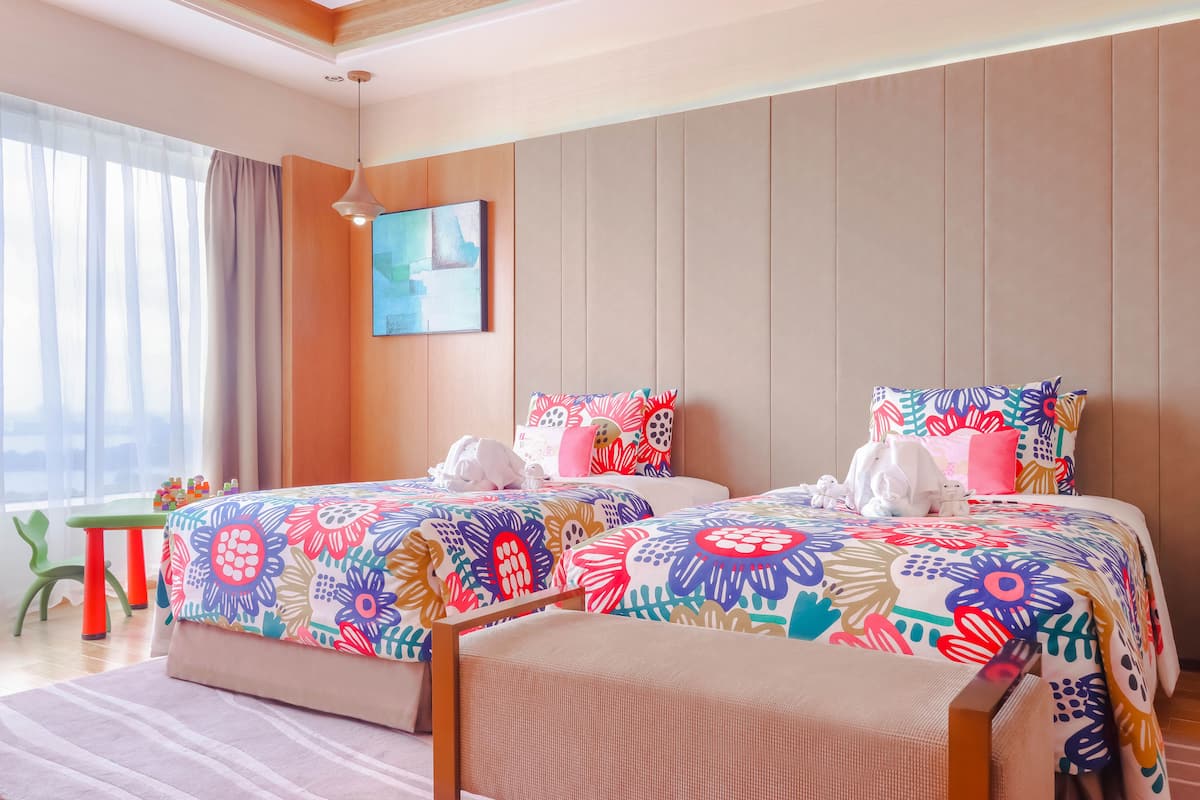 A hotel room with two beds, colorful floral bedding, a green chair, a small table, and a painting on the wall.
