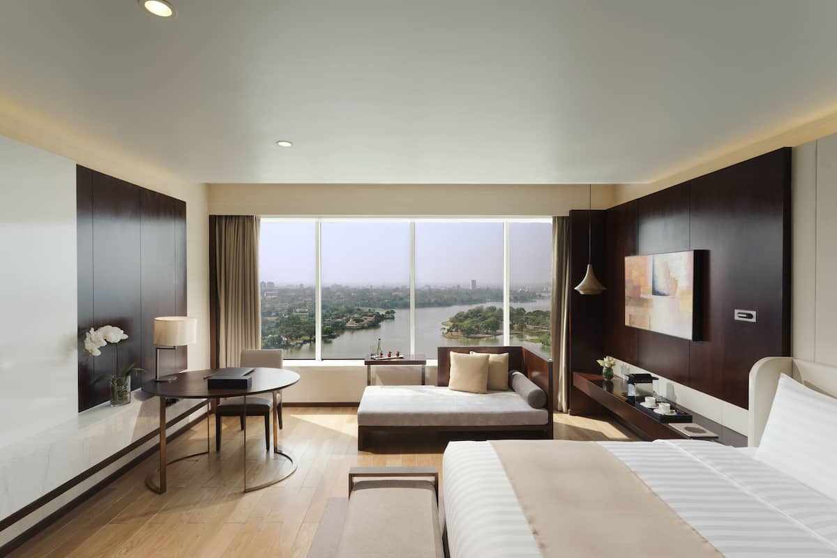 A modern hotel room with a large window, a bed, a desk, and a seating area.