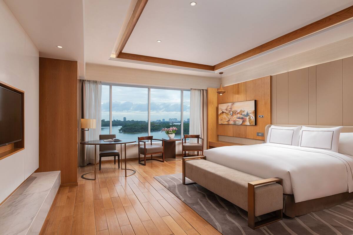 A modern hotel room with a large bed, a desk with chairs, a TV, and a view of a body of water through large windows.