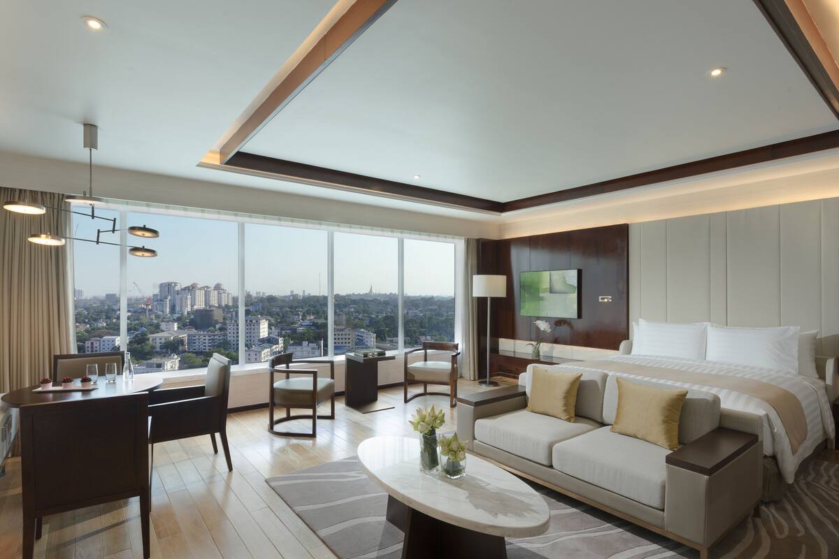 A modern hotel room with a large bed, a sofa, a dining area, and a view of the cityscape.