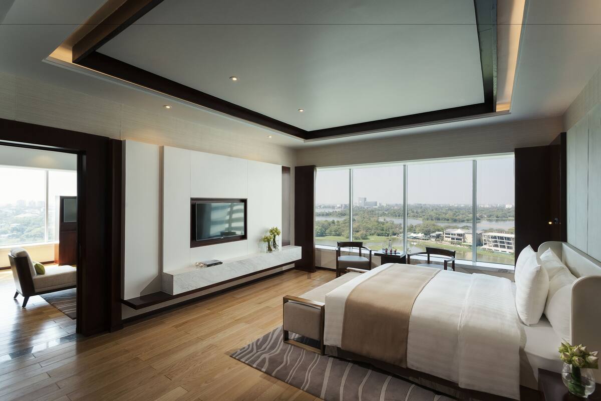 A modern hotel room with a large bed, a TV mounted on the wall, and a view of a cityscape through large windows.