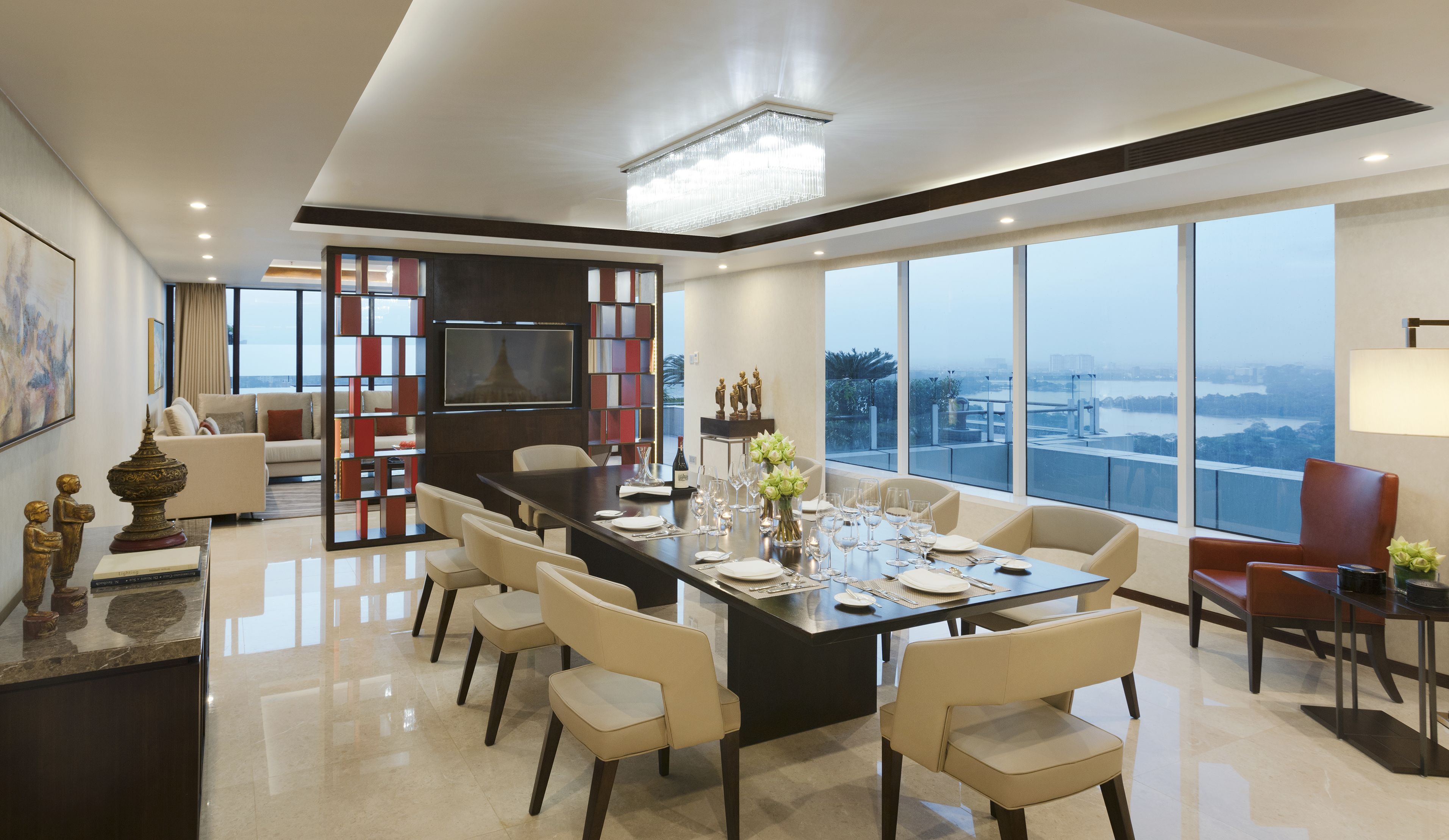 Un comedor moderno con una gran mesa dispuesta para seis personas, rodeada de sillas. Hay un televisor montado en la pared, una estantería y vistas de la ciudad a través de amplios ventanales.