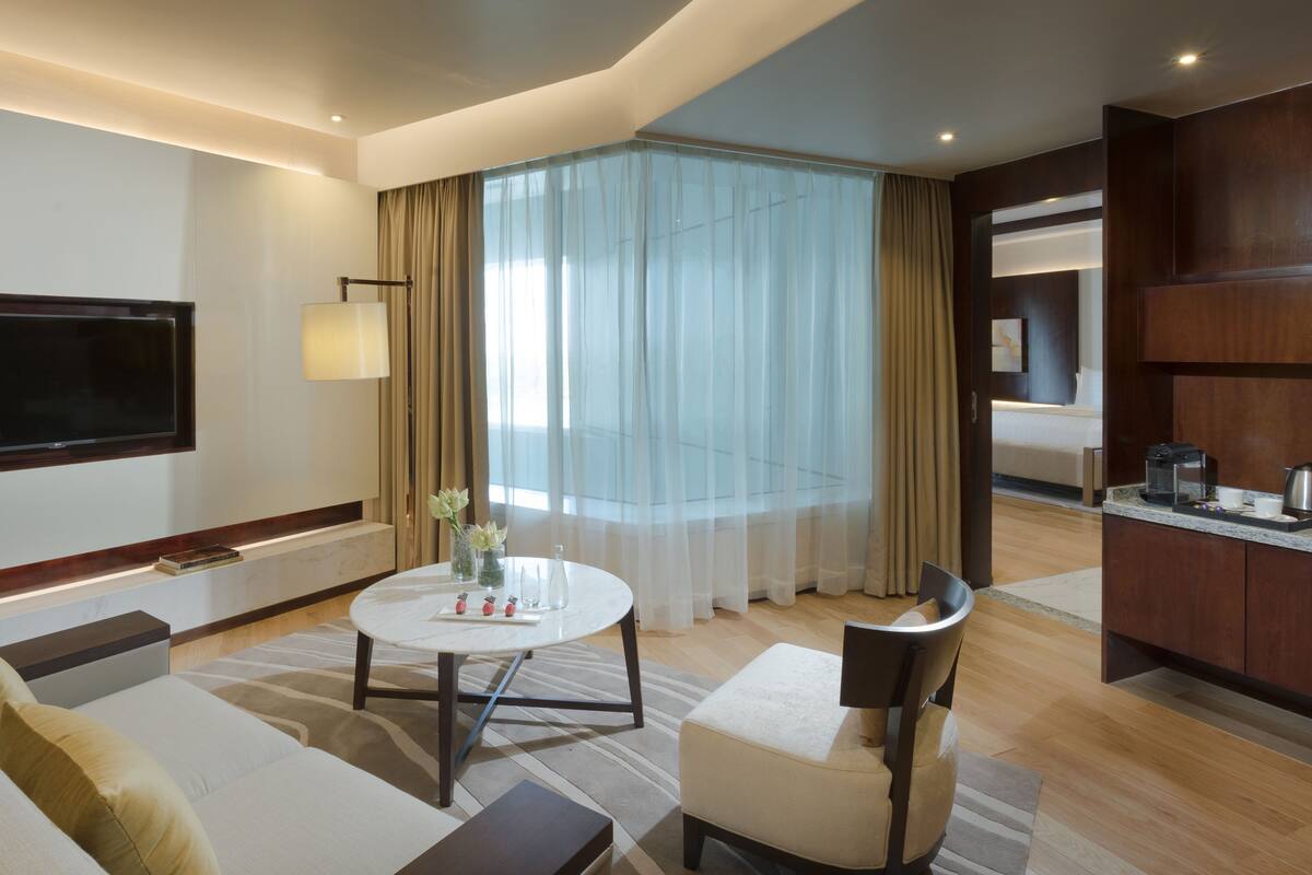 A modern hotel room with a flat-screen TV, a round dining table, a sofa, a chair, and a kitchen area.