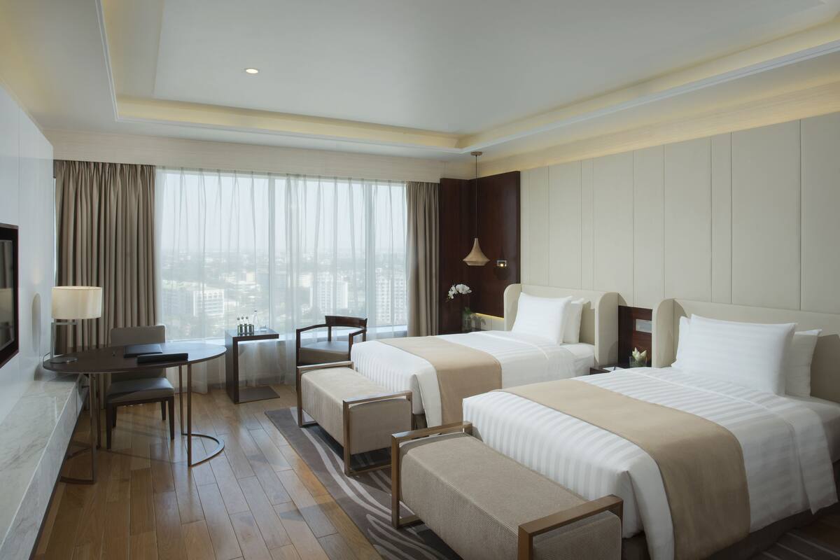 A hotel room with two beds, a desk, a chair, a TV, and a large window with curtains.