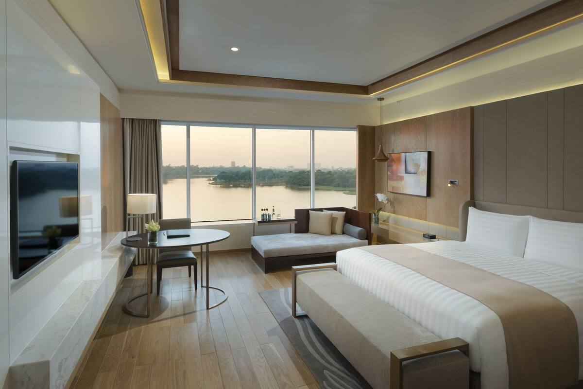A hotel room with a large bed, a sofa, a desk, and a TV. There is a large window with a view of a body of water and trees.