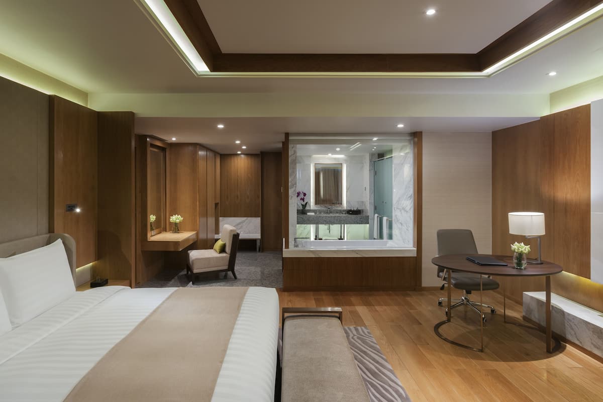 A hotel room with a large bed, a desk with a chair, a small seating area with a chair and a table, and a bathroom visible through a glass door.