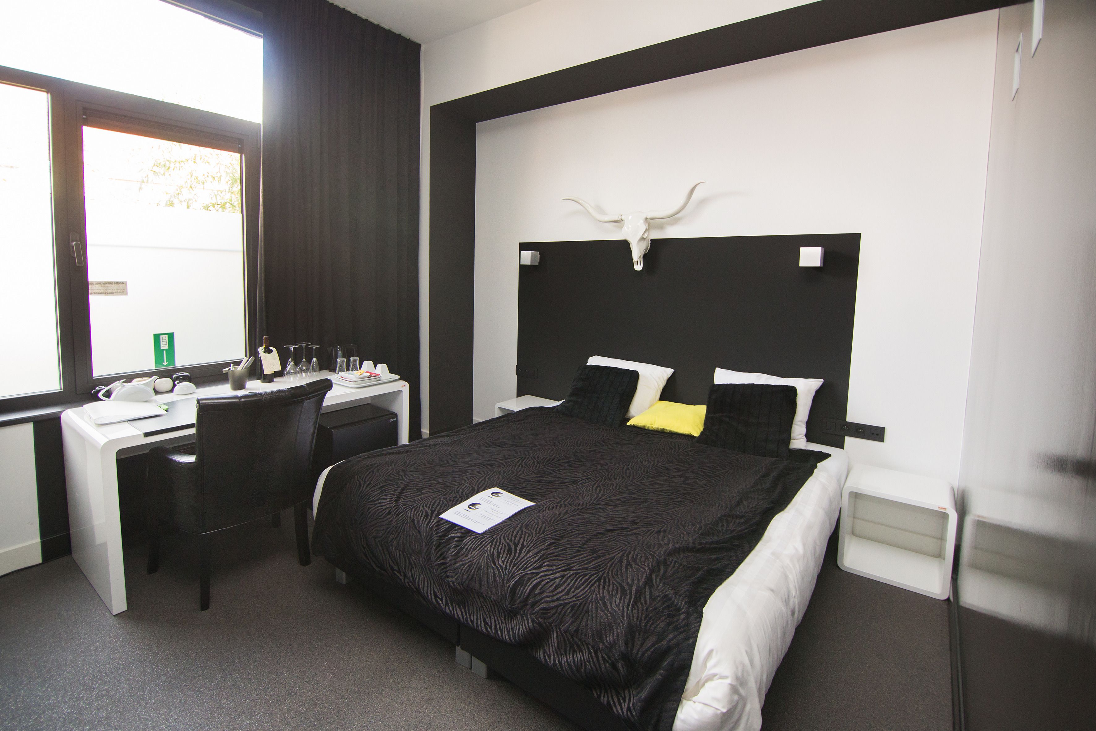 Una habitació d'hotel amb llit, escriptori i cadira. Hi ha una finestra amb cortines, una paret blanca amb capçal de llit negre i una paret blanca amb dibuixos en blanc i negre.