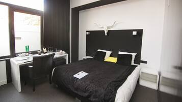 Una habitació d'hotel amb llit, escriptori i cadira. Hi ha una finestra amb cortines, una paret blanca amb capçal de llit negre i una paret blanca amb dibuixos en blanc i negre.