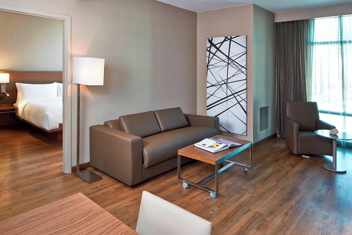 Una habitación de hotel moderna con sofá, una mesita, una silla, cama y una gran obra de arte abstracta en la pared.