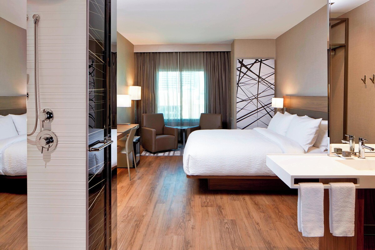 Habitación de hotel con una cama grande, un escritorio, una silla, ducha y baño con lavamanos y espejo.