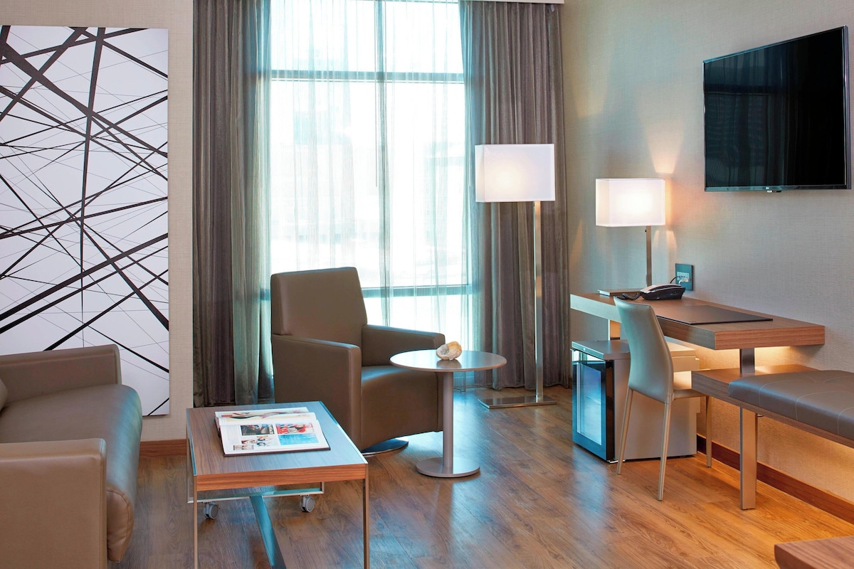Une chambre d’hôtel moderne, équipée d’un canapé, d’une petite table ronde, d’un bureau avec une chaise, d’une lampe et d’une grande fenêtre avec des rideaux.