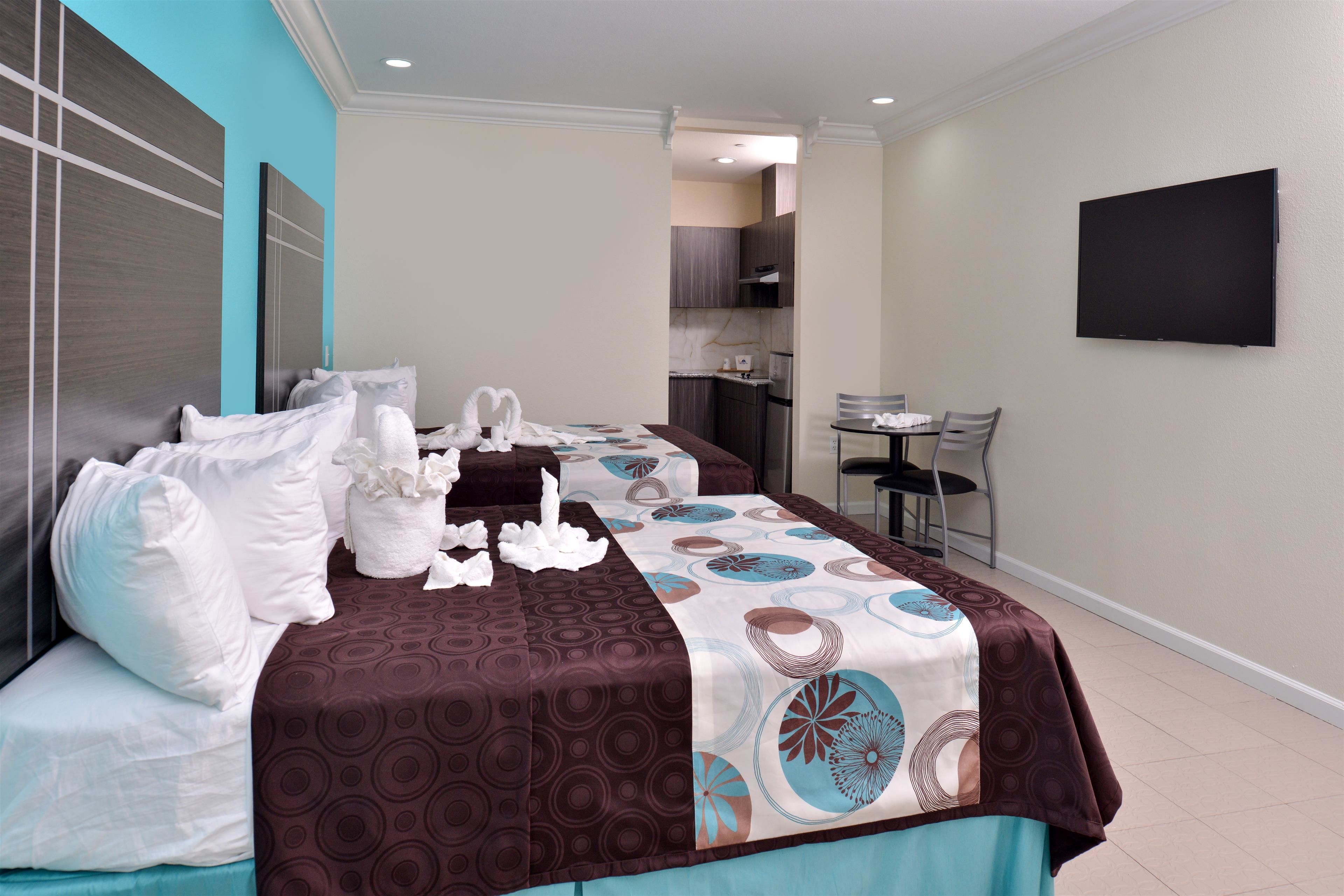 A hotel room with a bed, a desk, a chair, a flat-screen TV, and a kitchen area in the background.