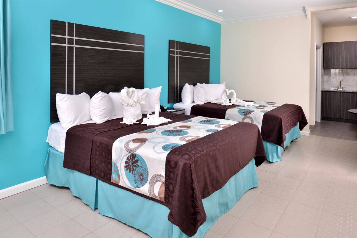 Une chambre d’hôtel avec deux lits, chacun doté d’une tête de lit, une salle de bain avec un lavabo et un miroir, et une petite kitchenette.