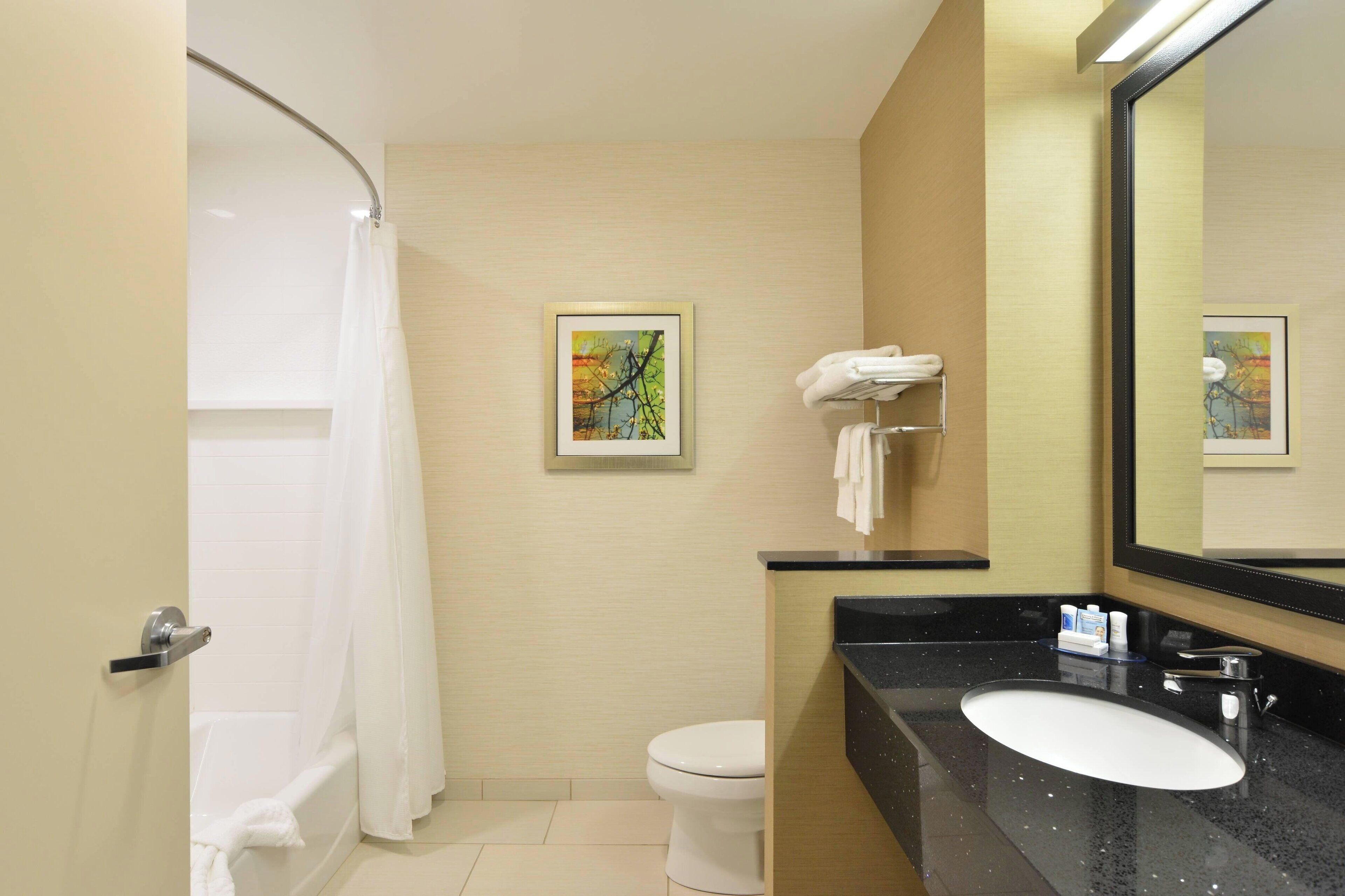 A modern bathroom with a bathtub, toilet, and sink. There is a framed picture on the wall and a towel rack with towels.