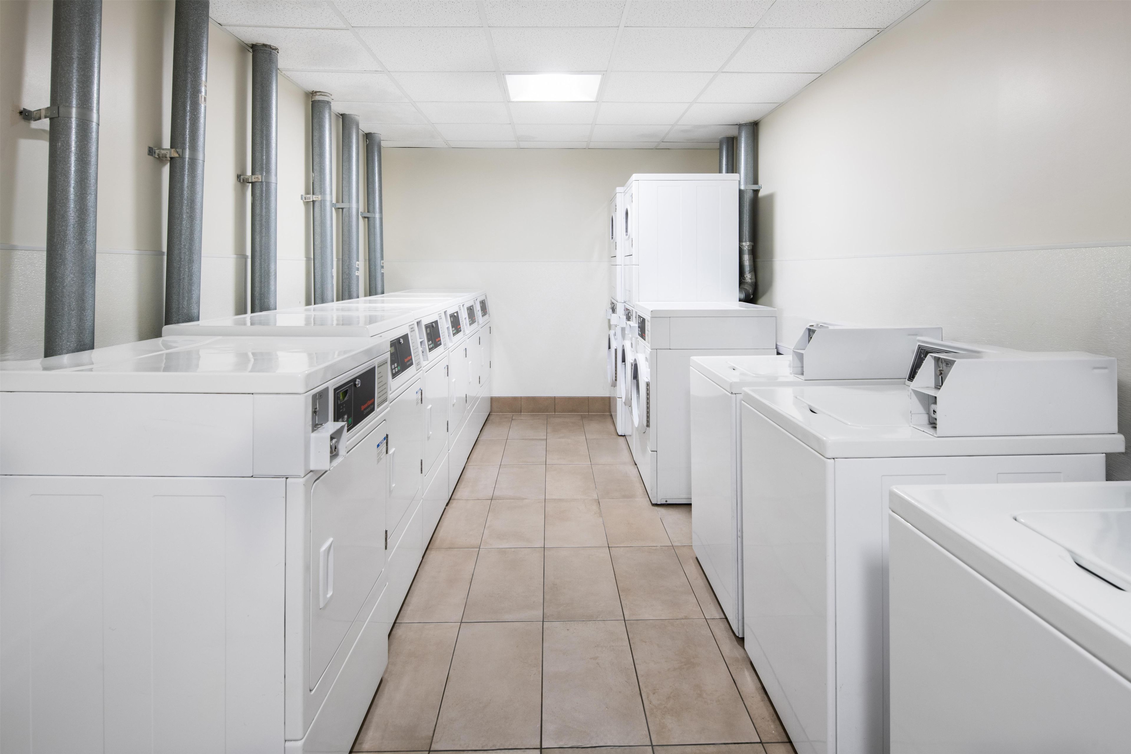 A laundry facility with rows of washing machines and dryers, a tiled floor, and a plain white wall.