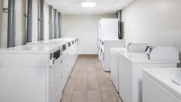 A laundry facility with rows of washing machines and dryers, a tiled floor, and a plain white wall.