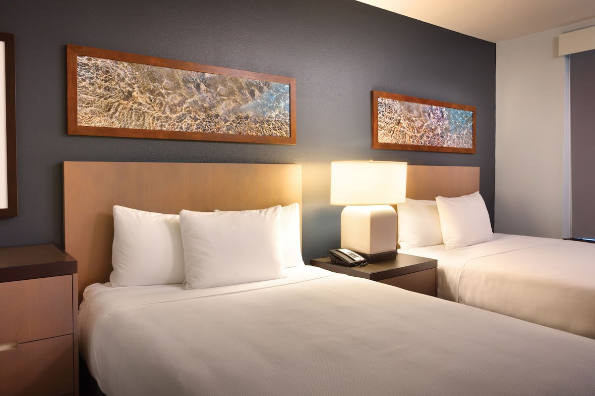 A hotel room with two beds, a nightstand with a lamp, and two framed abstract paintings on the wall.