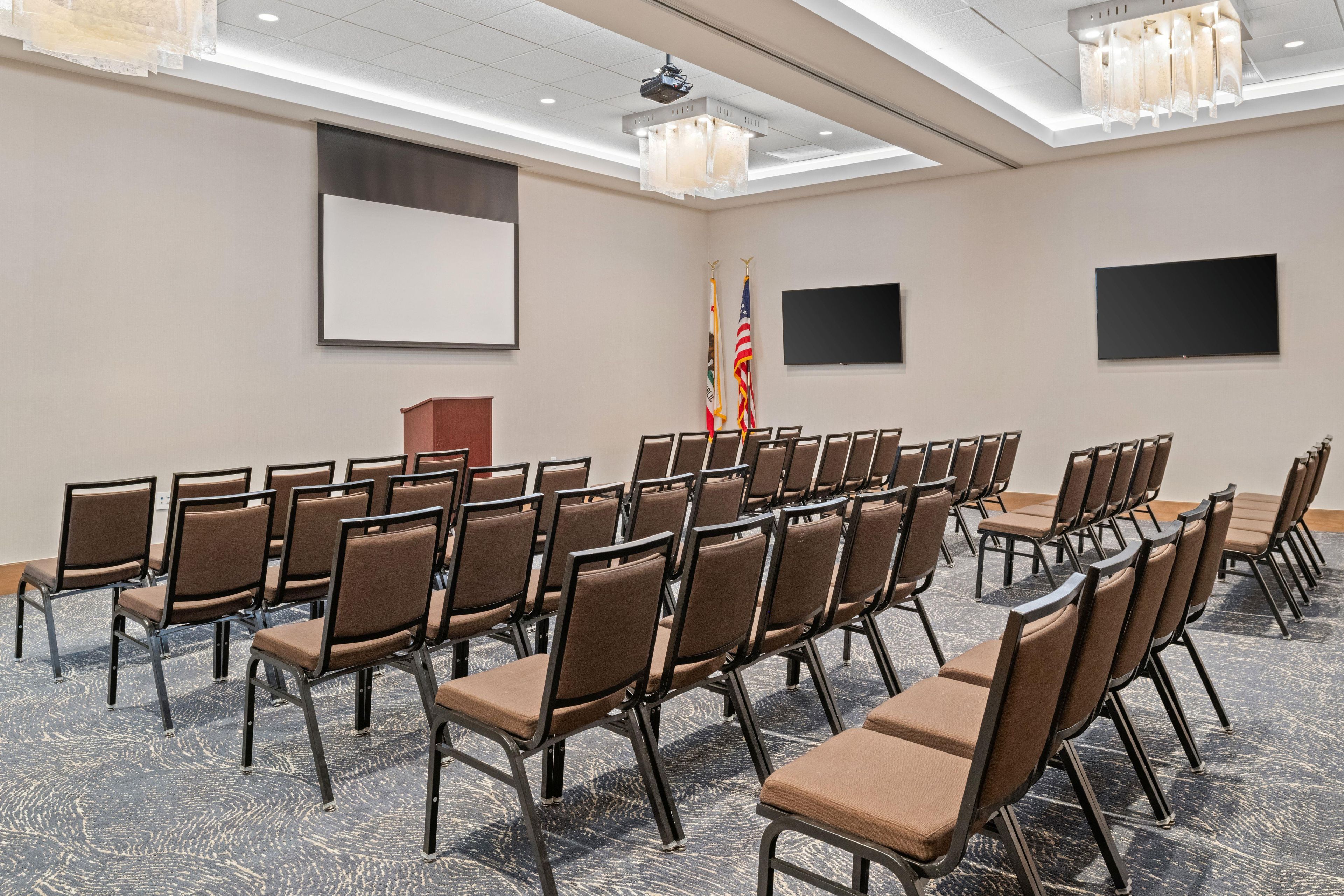 A conference room with rows of chairs, a podium, a projector screen, and two flags.