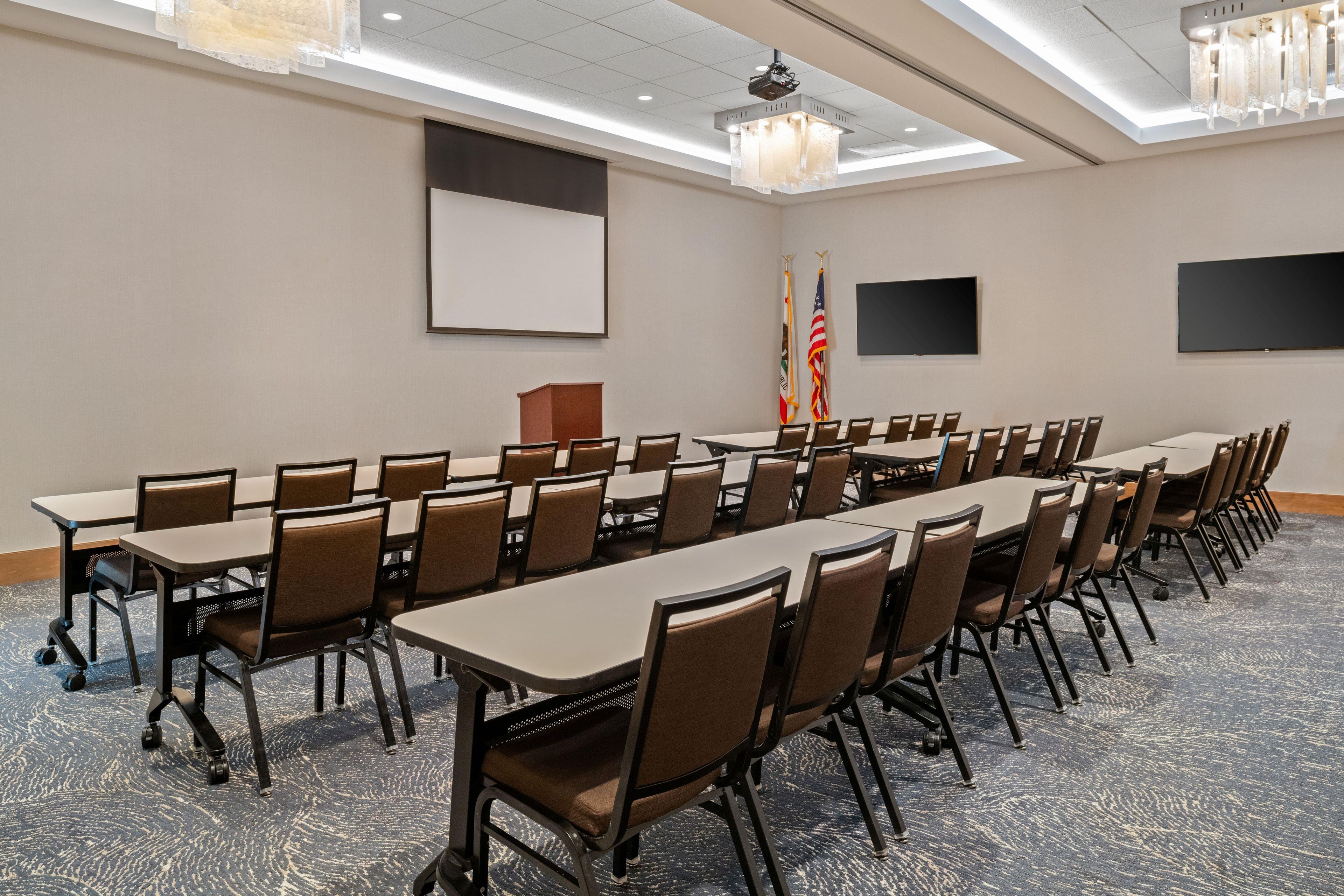 A conference room with long tables and chairs, flags, a podium, and a projector screen.