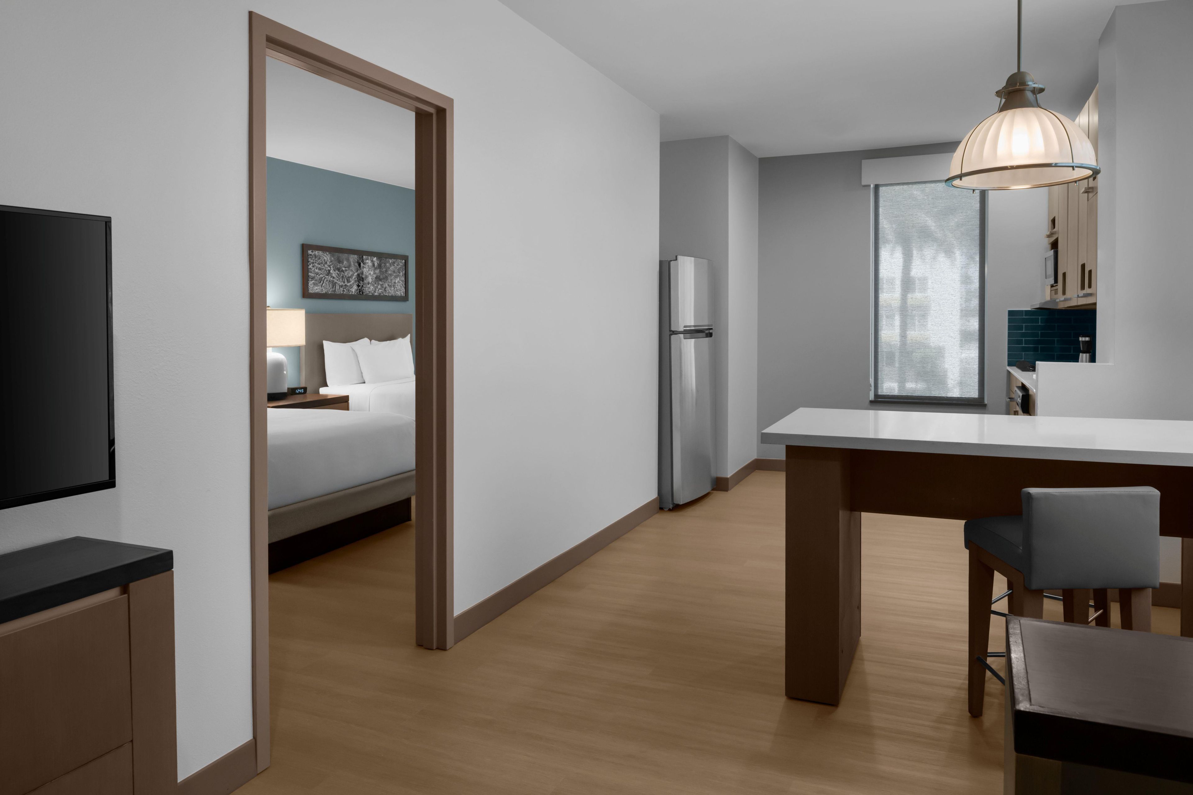 A modern hotel room with a bed, a desk, a refrigerator, a television, and a dining area with chairs.