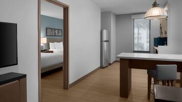 A modern hotel room with a bed, a desk, a refrigerator, a television, and a dining area with chairs.