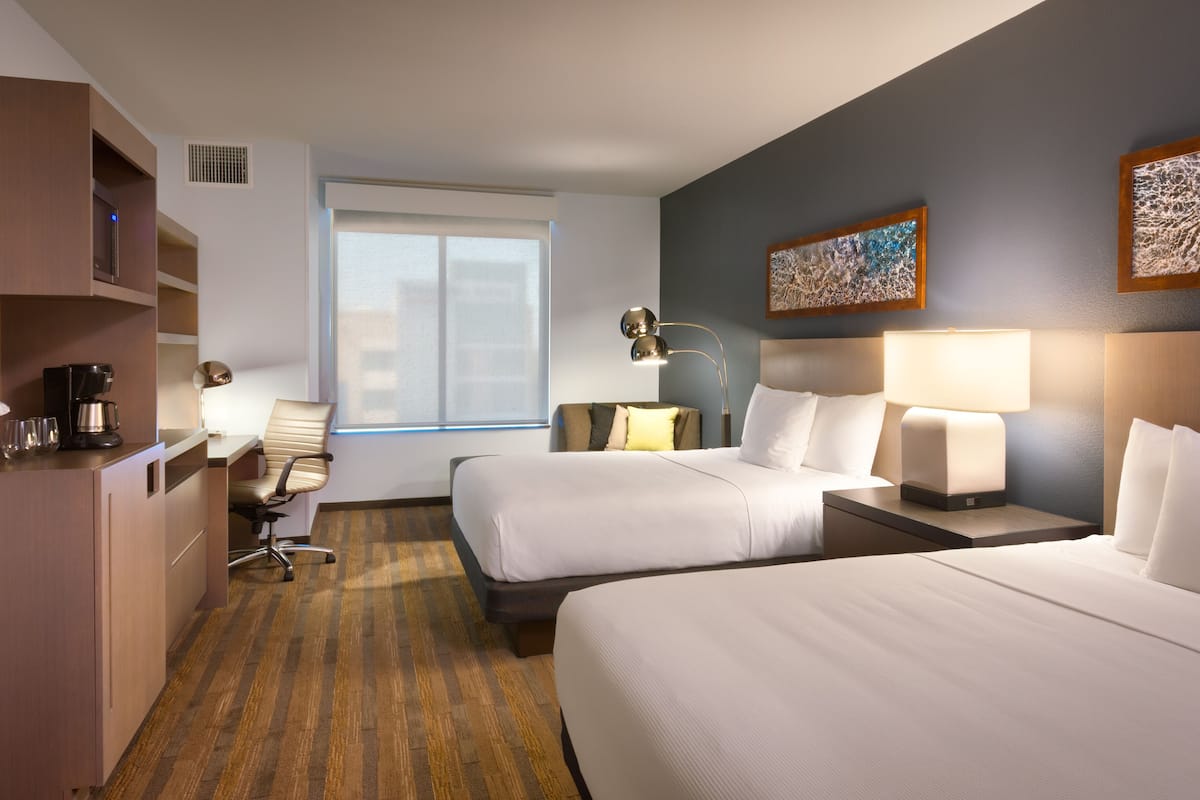 A hotel room with two beds, a desk, a chair, a coffee machine, and artwork on the walls.